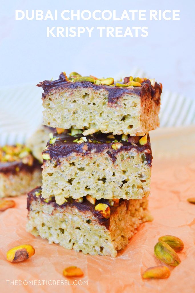 a trio stack of dubai-style rice krispy treats sits on a piece of brown parchment paper. there is a small scattering of shelled pistachios around the stack, and a striped dish cloth in the background.